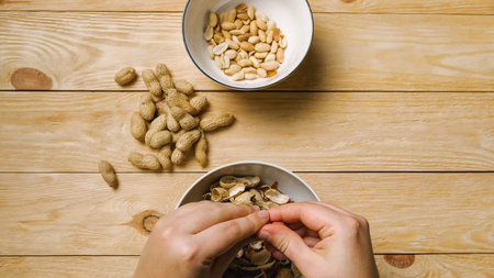 Close-up of women's hands peeling peanuts over bowls. A handful of peanuts lie on a wooden table....
