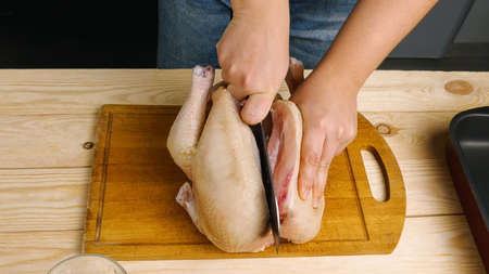 A woman is cutting a raw chicken carcass in half on a cutting board with a large knife. Cooking at homeの写真素材
