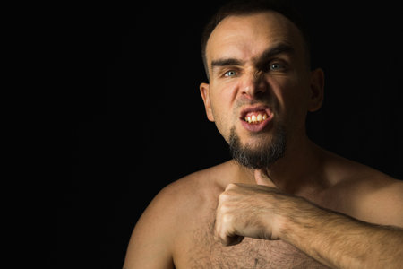 A man with an angry expression on his face, a human being threatening his throat with his thumb. Portrait of shirtless man with thumb at throat. black backgroundの写真素材