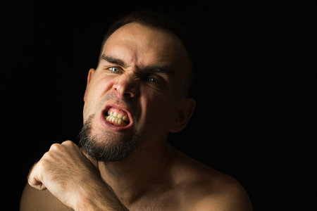 Shirtless bearded man, threatening with his thumb at his throat. Portrait of an aggressive man on a black background. The man grits his teeth, looking into the cameraの写真素材