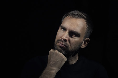 Close up portrait of a bearded man against a black background. Man resting his chin on his fist....