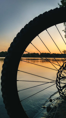 The silhouette of a mountain bike wheel on a river bank against the colorful backdrop of a sunset....
