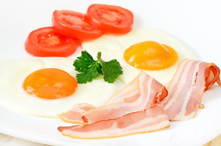 Breakfast with bacon, fried eggs and slices tomatoes on white plateの写真素材