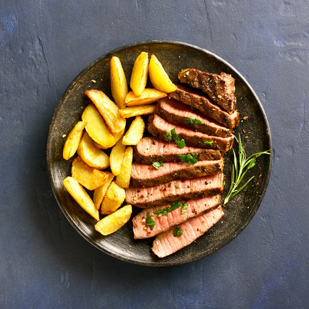 Sliced medium rare roast beef with potato wedges on plate over stone background. Top view, flat layの写真素材