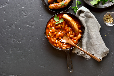 White beans in tomato sauce and grilled sausages in frying pan over black stone background with copy space. Top view, flat layの写真素材