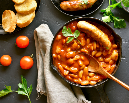 Close up of baked white beans in tomato sauce and grilled sausages in frying pan over black stone background. Top view, flat layの写真素材