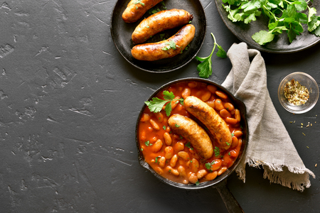 Tasty grilled sausages with baked white beans in tomato sauce in frying pan over black stone background with copy space. Top view, flat layの写真素材