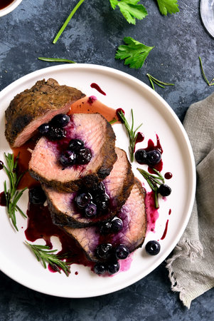 Roast beef with berry sauce. Sliced grilled beef with blueberry sauce on white plate over blue stone background. Top view, flat layの写真素材