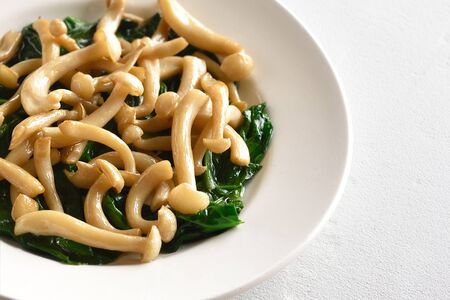 Close up of stir-fried white beech mushrooms with leaves of spinach on white stone backgroundの写真素材
