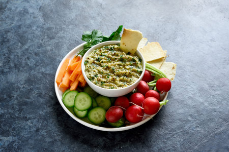 Baba ganoush (roasted eggplant dip) in bowl and fresh organic vegetables over blue stone background...