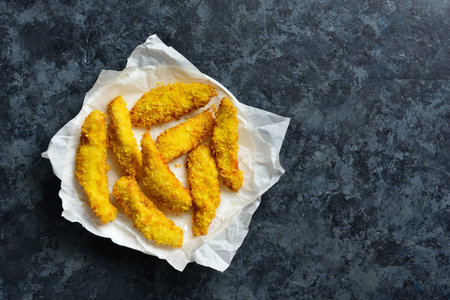 Crispy fried breaded chicken breast strips on paper over blue stone background with free space....