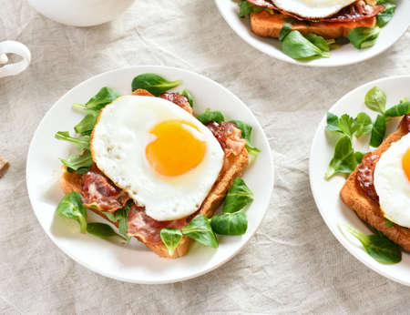 Close up view of open sandwiches with fried eggs, bacon and vegetable leaves on plate over light background.の写真素材