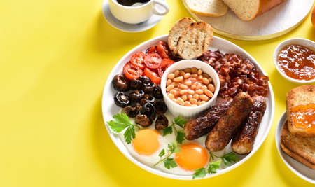 Full english breakfast with fried egg, bacon, sausage, beans and mushrooms on yellow background with copy space.の写真素材
