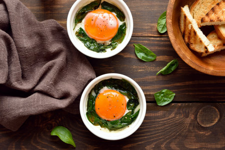 Eggs en cocotte (baked eggs) in ramekins with cream, cheese and spinach on wooden background. Top view, flat layの写真素材