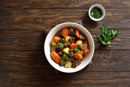 Beef meat stewed with potatoes, carrots and spices in white bowl over wooden background. Comfort healthy food for dinner. Top view, flat layの写真素材