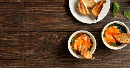 Eggs en cocotte (baked eggs) in ramekins with cream, cheese and spinach on wooden background with free space. Served with warm toast for dipping. Top view, flat layの写真素材