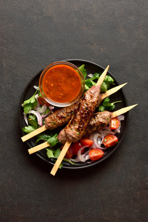 Grilled lula kebab on wooden skewers served with fresh vegetables and tomato sauce on black plate on dark background with free space. Top view, flat lay, closeupの写真素材