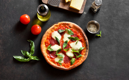 Pizza with mozzarella, pepperoni, tomato sauce and basil. Homemade pizza with tomatoes, cheese, sausage on dark background. Top view, flat lay.の写真素材