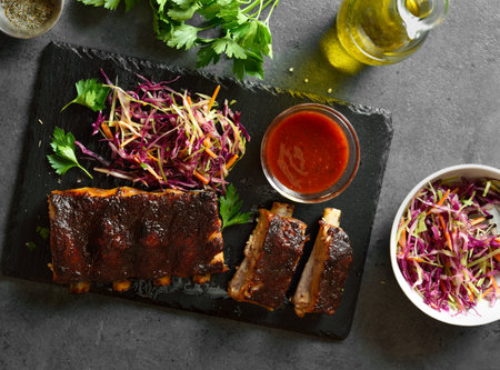 Spicy grilled spare ribs served with fresh cabbage salad and sauce on slate platter over dark stone background. Top view, flat layの写真素材