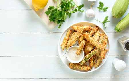 Baked crispy zucchini fries served with dipping sauce on plate over light wooden background with free space for text. Top view, flat layの写真素材