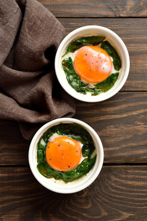 Diet meal for breakfast Eggs en cocotte (baked eggs) in ramekins with cream, cheese and spinach on wooden background. Top view, flat layの写真素材