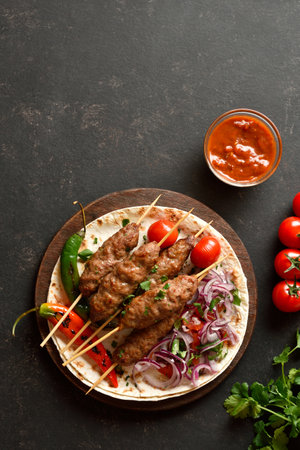 Grilled lula kebab with fresh vegetables on flat bread on wooden platter on black background. Top view, flat layの写真素材