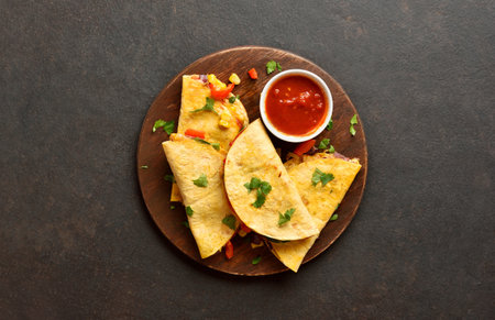 Delicious quesadilla with fresh vegetables served with tomato sauce on wooden platter on brown stone background. Top view, flat layの写真素材