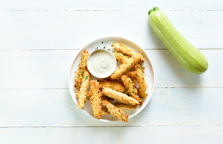 Baked crispy zucchini fries with dipping sauce on plate over light wooden background. Top view, flat layの写真素材