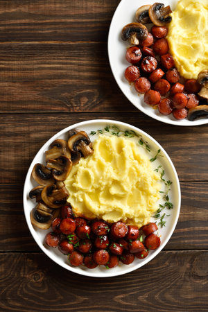 Mashed potatoes with fried sweet and spicy sausage bites and mushrooms on white plate on brown wooden background. Top view, flat layの写真素材