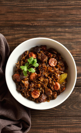 Stewed cabbage (polish bigos) with sauerkraut, mushrooms, smoked meats and spices in bowl over wooden background. Top view, flat layの写真素材