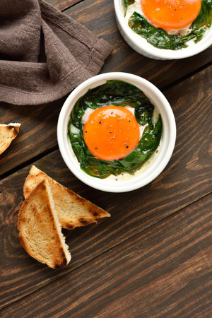 Eggs en cocotte (baked eggs) in ramekins with cream, cheese and spinach on wooden table. Top view, flat layの写真素材