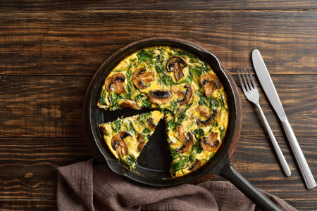 Frittata with spinach and champignon mushrooms in cast iron skillet on brown wooden background. Top...