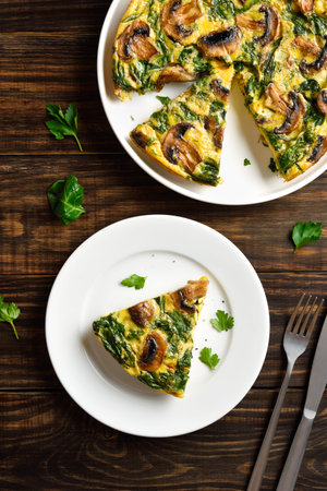 Sliced frittata with spinach and champignon mushrooms on white plate on brown wooden table. Top...