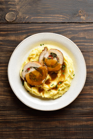 Pork rolls with mushrooms, cheese, red bell pepper in gravy sauce served with mashed potatoes on light plate on brown wooden background. Top view, flat layの写真素材