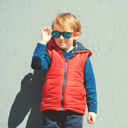 Stylish kid in trendy sunglasses. Kids fashion. Cute little blondy boy in red jacket standing over...