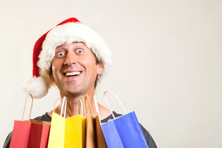 Surprised man in Christmas hat holds shopping bags, isolated on white. Christmas shopping and sales concept. Christmas discounts. Funny man shopper in santa hat holding gifts in shopping bagsの写真素材