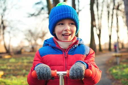 Cute little boy outdoors portrait. Happy child in warm clothes is riding scooter at autumn park. Happy and healthy childhood. Autumn holiday. Fashionable little boy having fun outdoors in autumnの写真素材