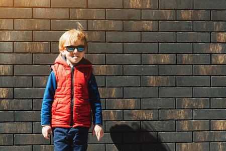 Cute stylish boy against brick wall outside. Blond fashionable boy in sunglasses, wearing red vest, looking to camera and smiling. Boy posing in sunny day. Kids fashion. Trendy kids sunglassesの写真素材