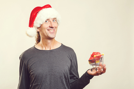 Christmas sales concept. Happy Christmas man holding small shopping cart with christmas gift, isolated. Copy space. New Year's Holiday. Winter sales. Smiling man in Santa hat with shopping basketの写真素材