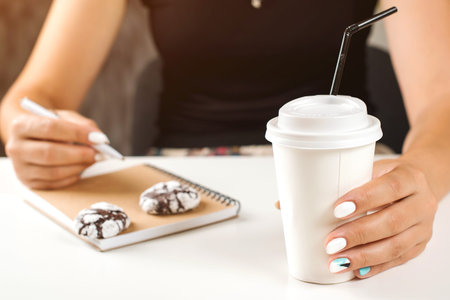 Young woman drinking coffee from paper cup. Morning coffee and planning your day at notebook. Woman holds a disposable coffee cupの写真素材