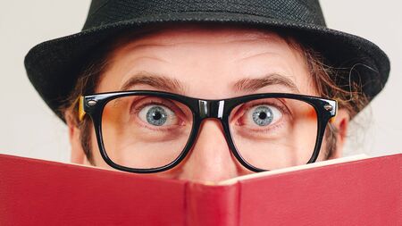 Excited young nerd guy holding book. Nerd in glasses. Funny man looking to camera. Never too late to study. Amazing man has great idea. Male nerd hiding behind books. Nerd hipster guy in hatの写真素材