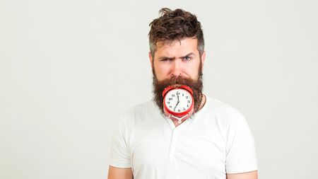 Handsome man holds the clock with his mouth. Crazy bearded hipster with clock. Brutal bearded guy with alarm clock on white background. People and time conceptの写真素材