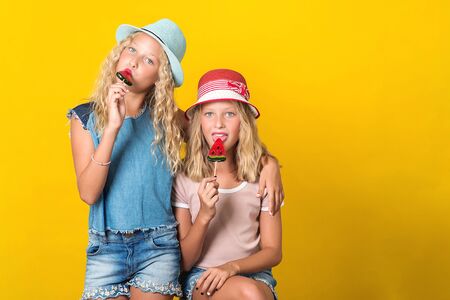 Happy twin sisters in summer hats. Teenagers girls posing on yellow background. Sisters in stylish summer outfit. Funny girls holding lolilops. Summer holidays concept. Copy spaceの写真素材