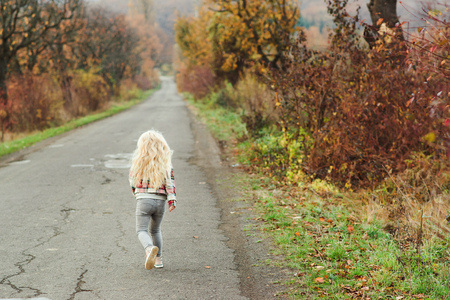 Happy little girl with blonde long hair running away on the road, back view. Walk in autumn time. Stylish fashion child outdoors. Autumn holidays concept. Childhood, leisure and peopleの写真素材