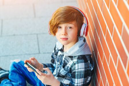 Cute redhead boy with stylish hairstyle. School boy listen to music in headphones. Fashion teenager at city walk. A handsome boy in a plaid shirt. Kid plays games on phoneの写真素材