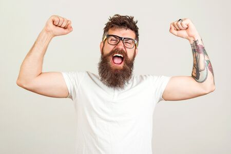 Screaming bearded brutal man with hands up. Excited bearded man in glasses celebrating success. Goal, Winner, Celebrating. Face expression. Bearded hipster with tattooの写真素材