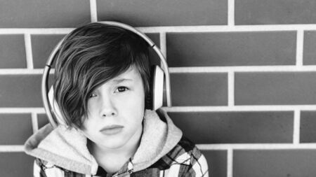 Stylish young boy in headphones listening to music over brick wall. Teenages and emotions concept. Sad handsome boy teen with headphones looking to cameraの写真素材