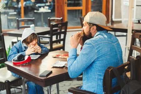 Father and son eating pizza at a pizzeria outdoors. Hungry schoolboy with father has a lunch at cafe. Stylish family spending time in pizzeria. Delicious pizza for lunch.の写真素材