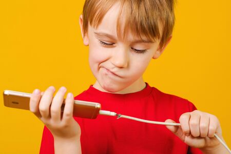 Boy holding a mobile phone with a defective charging cord. Broken smartphone, defect of charger cable. Boy gesturing shock and disorder. Boy thinks how to repair cabel.の写真素材