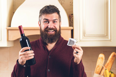 Handsome man with a bottle of wine and a condom. Bearded guy getting ready for a date. Man holding condom. Safety sex concept. Valentine's holiday. Bearded guy keeps contraceptive, advertises.の写真素材